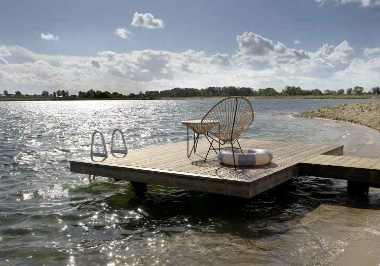 Holzsteg mit Stuhl, Handtuch und Schwimmreifen am Ufer des Sees, Sonnenlicht spiegelt sich im Wasser.