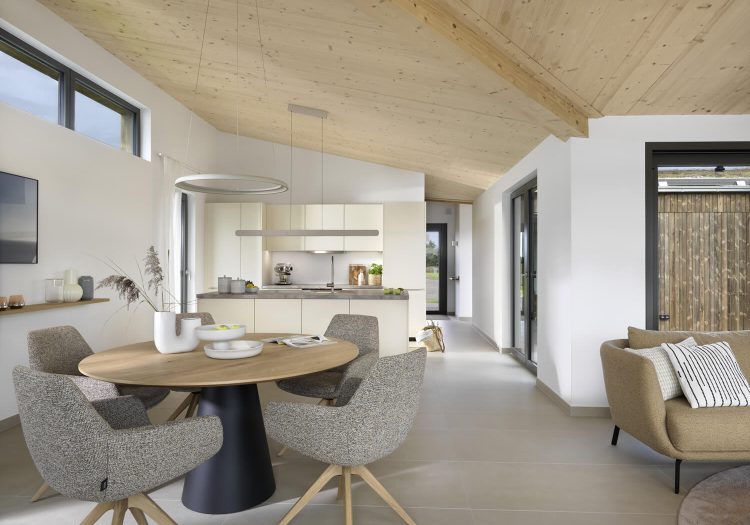 Helles Esszimmer mit Rundtisch, offenen Küchenbereich und großen Fenstern.